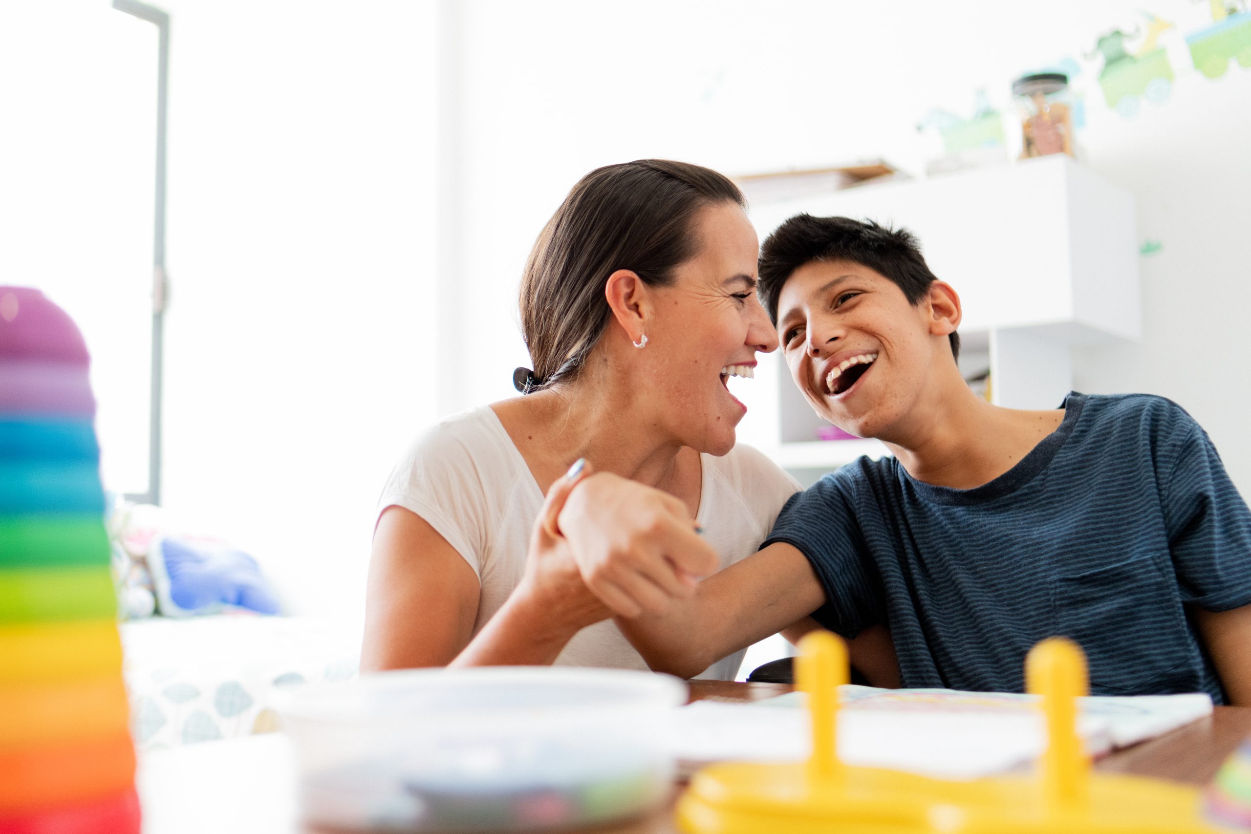 image: a woman and a kid with obvious disabilities, both with dark skin and dark hair in a house setting laughing and holding each other