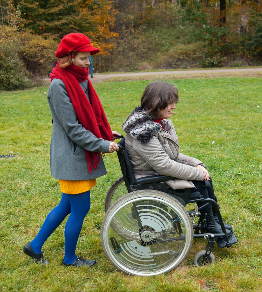 Young woman pushes another woman in a wheelchair through a field on a fall day.