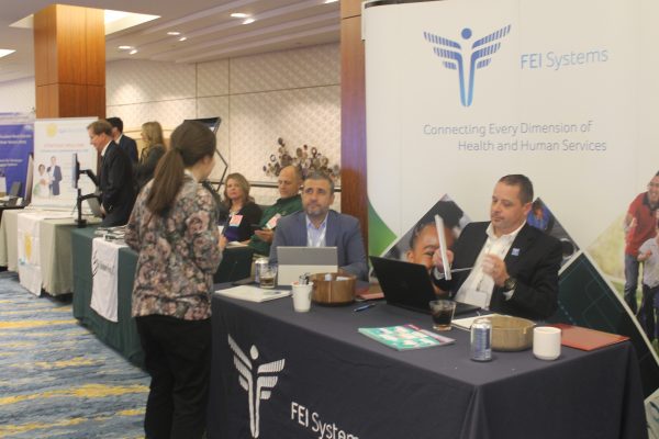 image: multiple diverse people in a conference setting with two men with light skin and dark hair representing FEI Systems giving a talk to a woman in a conference setting