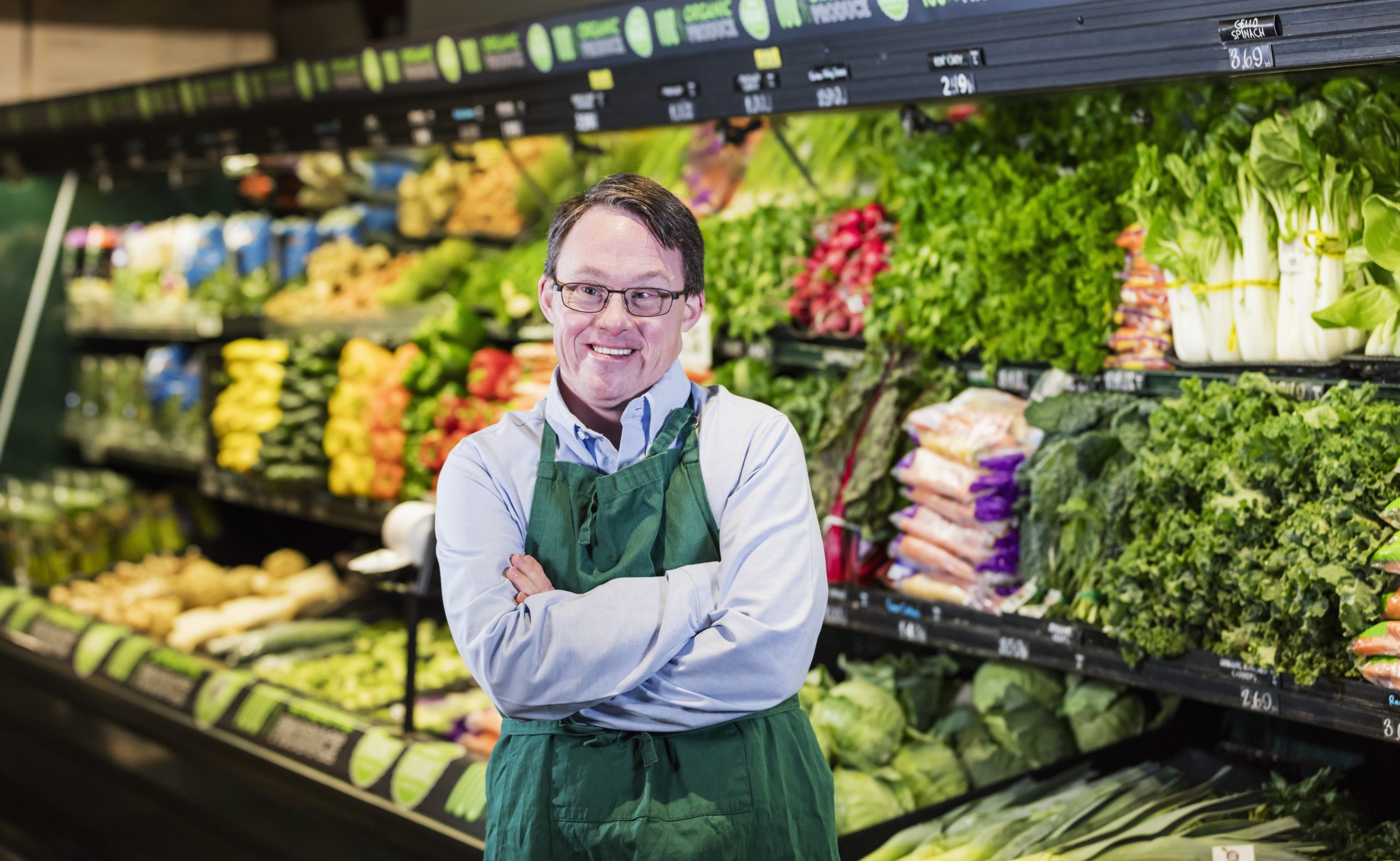 image: a man with light skin and dark hair, with obvious disabilities wearing a formal shirt and an apron in front of a vegetable wall in a market setting