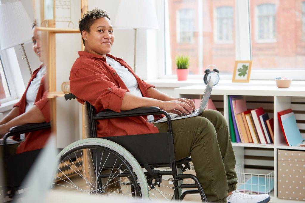 image: a woman using a wheelchair with dark skin and hair in casual attire, working on her laptop