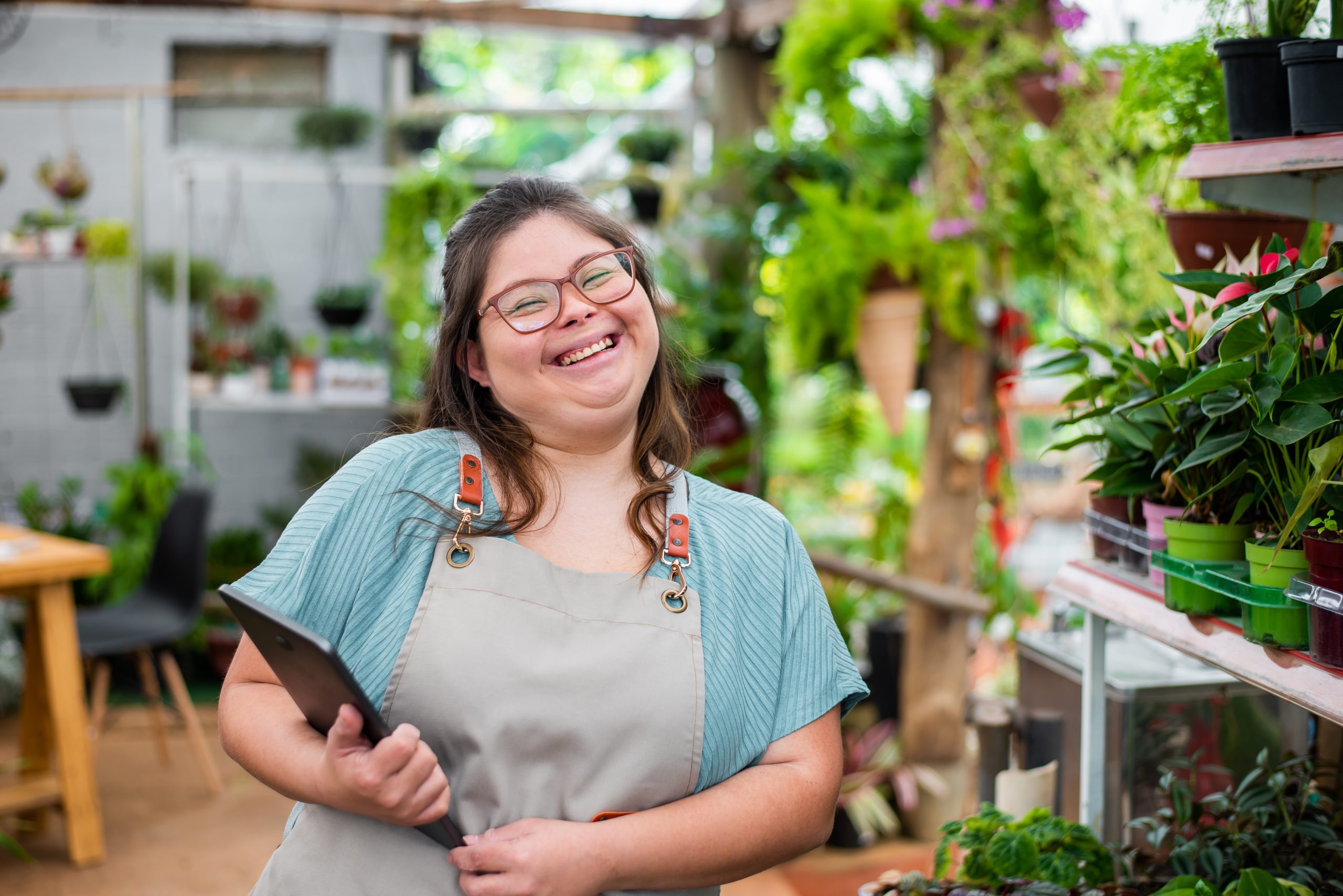 image: a woman with obvious disabilities with light skin and dark hair wearing an apron, holding a tablet in a garden shop setting smiling at the camera