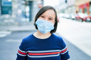 Image: a light skin and dark hair woman with obvious disabilities in a blue and red shirt wearing a face mask looking at the camera