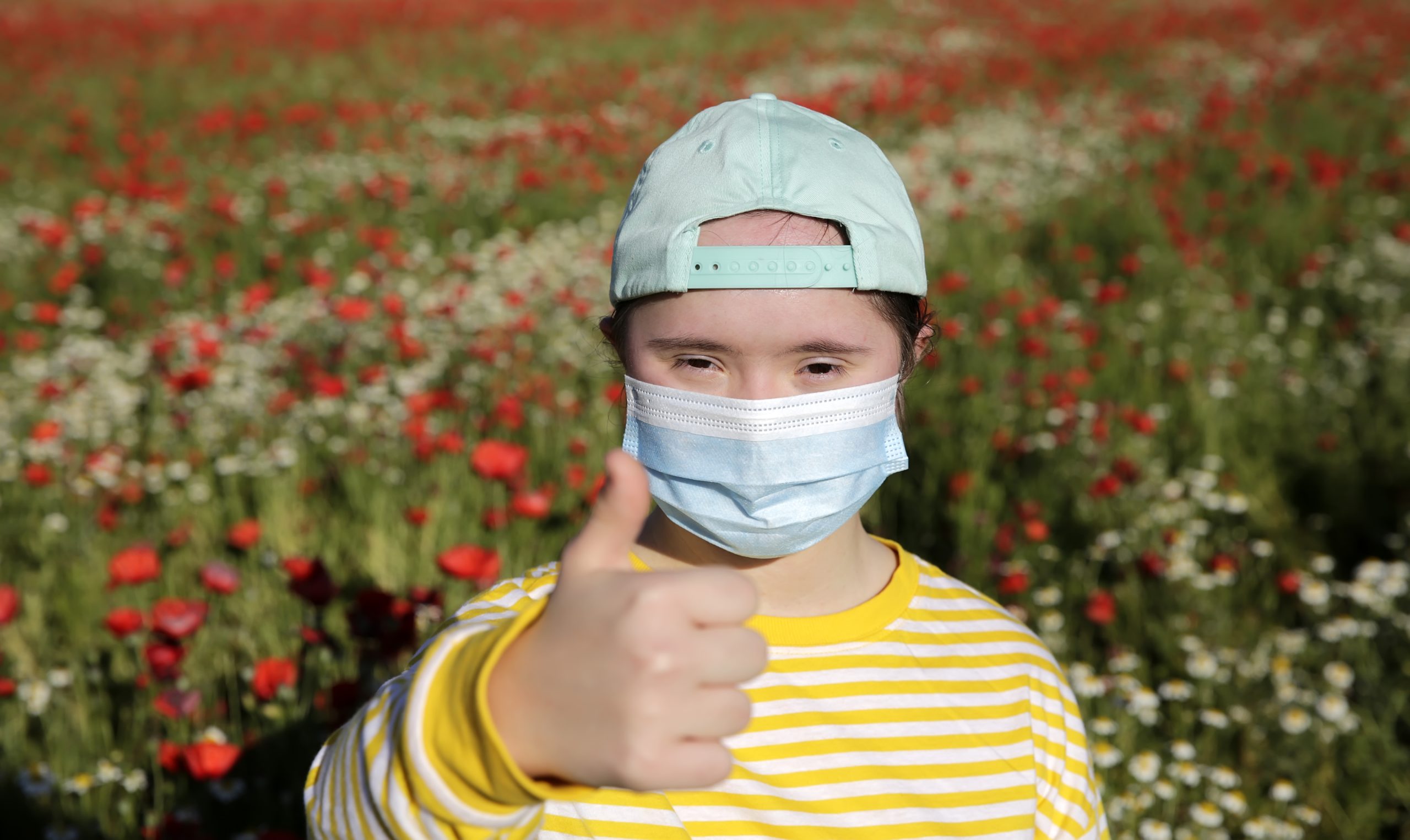 image: a girl with obvious disabilities wearing a yellow shirt, a backwards cap and a face mask holding a thumb up for the camera