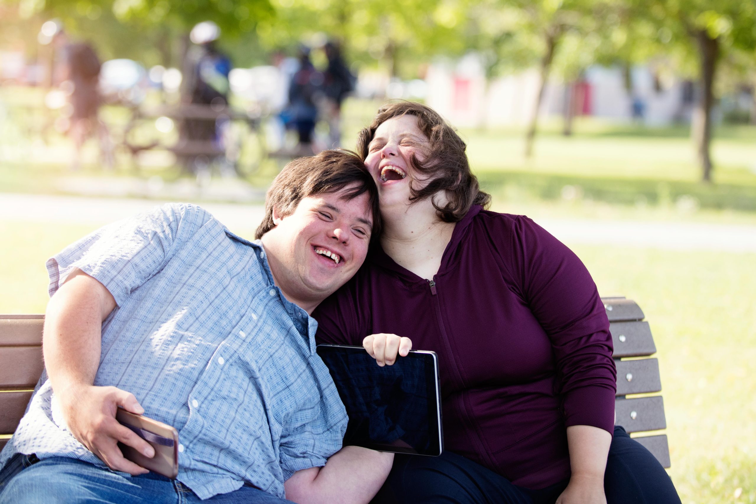 image: a man and a woman with light skin and dark hair, both with obvious disabilities in casual attire laughing in a park setting sitting in a bench
