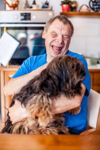 image: a man with light skin and light dark hair, with obvious disabilities in a blue shirt smiling/laughing holding a dog with dark hair