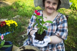 Image: a woman with light skin, dark hair and obvious disabilities in a garden setting holding a plant with a red flower