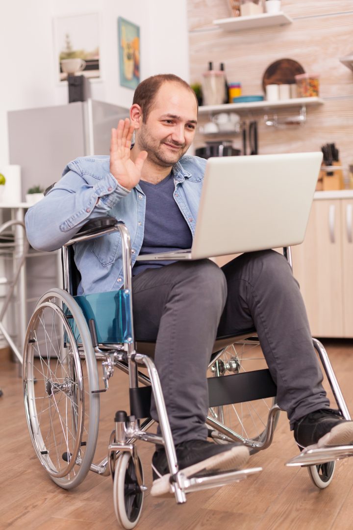 Image: a man wearing casual attire using a wheelchair waving at a laptop on his lap